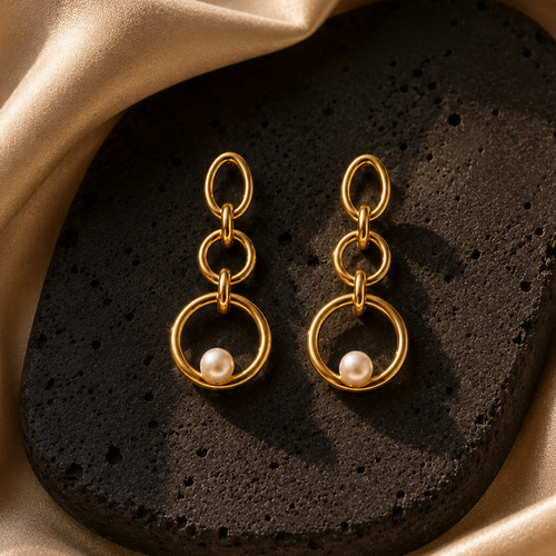 A pair of gold dangle earrings with three interlocking rings and a small white pearl at the bottom, displayed on a dark textured stone against a draped champagne silk background.