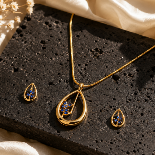 Gold jewelry set featuring a teardrop pendant necklace and matching drop earrings, both with blue gemstone accents, displayed on a black textured stone slab with cream satin fabric and dried flowers.